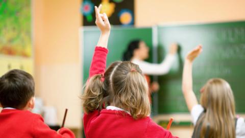 A classroom. A schoolgirl in a red cardigan raises her hand to answer a teacher's question. The teacher writes on a board in the background.