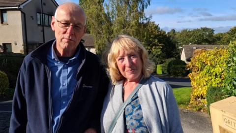 A man and woman look into the camera with neutral expressions on their faces. Behind them are a few houses surrounded by hedges and trees.