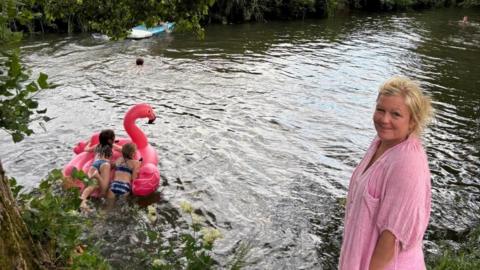 To the right of the image, Harriet Symon stands smiling at the camera, wearing a pink top. Behind her two young girls lie on their front on a pink inflatable flamingo on the river. 
