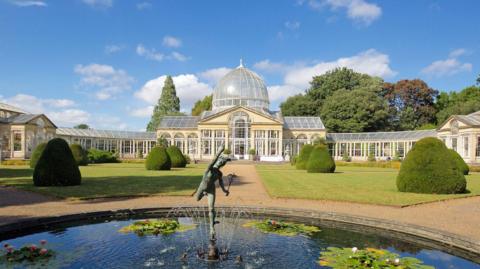 Neoclassical mansion with glass dome roof, and gardens with pond, as pictured in sunshine.