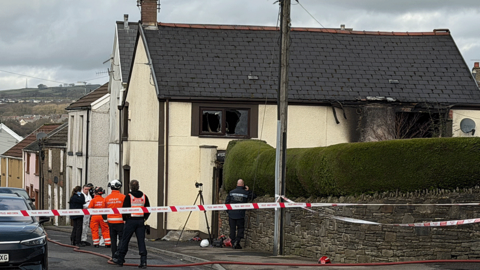 Investigators gather in the street by a house where first-floor windows have been either shattered or are missing and the yellow painted external walls have been partially blackened as if by fire damage