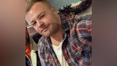 Selfie of Ben Crook. He wears a white t-shirt and a red and navy flannel overshirt. He smiles at the camera and has short blonde hair. 