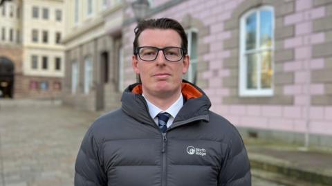 A man wearing glasses, a ties and a warm puffer coat stands upright outside and looks into the camera. 