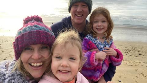 Alison Robinson grins at the camera, her arm around one of her young daughters. Alison is wearing a pink hat and grey scarf. She has blonde hair. Her daughter has blond hair and is wearing a pink jumper, she is grinning at the camera. Behind them is Alison's partner who is wearing a blue and green hat and a blue hoodie. He has his arm around their second daughter who has brown hair and is wearing a floral blue top and a pink and purple knitted jumper. The family are taking the selfie on the beach. 