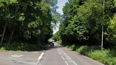 A road that has grass and trees on either side as well as a junction on the left. Other vehicles can be seen travelling forwards and a lamp-post is visible on the right hand side.