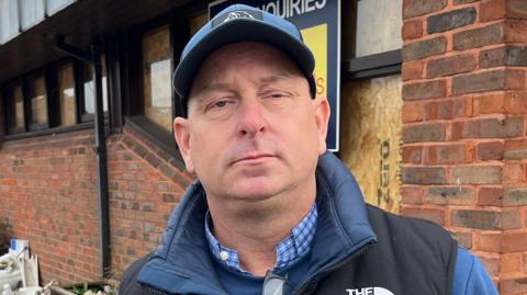 A man wearing a blue baseball cap, a blue body warmer over a blue top and a blue and white chequered shirt underneath. He is standing in front of a corner of an old brick pub with a large pile of debris and rubble to the left.
