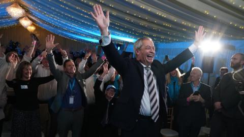 Reform UK leader Nigel Farage raising his hands in the air and smiling in a organza-clad tent with fairy lights on the ceiling. Lots of supporters are behind him also raising their hands. 