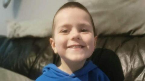 Kyran Durnin - a young boy with short, dark hair smiles at the camera while he sits on a dark, leather sofa. He is wearing a blue hoodie.