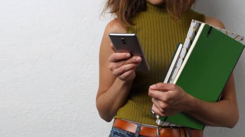 A stock image shows a woman from the chin down wearing a yellow sleeveless top and jeans. She is holding several folders and notepads under her left arm and a phone in her right hand, which she appears to be locking at. She is standing in front of a white textured wall.