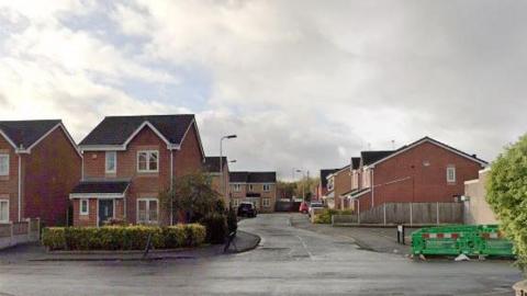 The entrance to Boxwood Gardens in St Helens, a house is to the left and roadworks sit on the edge of the path on the right, the camera looks down the estate.