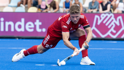 A man wearing a mainly red top, shorts and socks is playing hockey, holding a stick with the ball next to it on a blue surface. People in the crowd are blurred in the background.