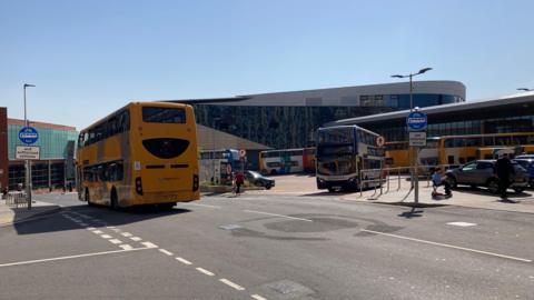 Buses - one yellow and another blue and white - using a bus station on a sunny day while people walking on the neighbouring streets.