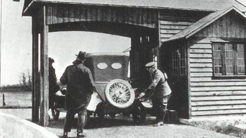 A black and white image shows a uniformed AA patrolman wearing a cap filling a car with fuel. The car sits under a wooden canopy next to a wooden single-storey building with a window. To the side of the car a man stands with his back to the camera. He is wearing a long dark coat over dark trouser and a dark hat with a brim. A third man wearing a flat-cap style hat can be seen to the rear of the wooden canopy, on the left side behind the front end of the car.
