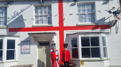 Two people dressed as an English knight and a Beefeater stood outside a white pub with a red stripe which makes it look like a English flag.