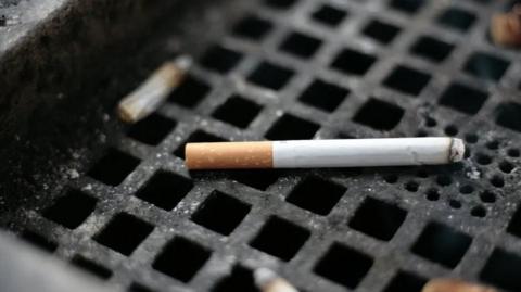 A white cigarette, with a brown filter, has just been lit and lies on the black grid of an ashtray. There's a smoked butt  and ash nearby.