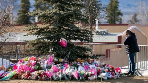 A woman stands beside a memorial for the victims of the Tumbler Ridge shooting, set up around a spruce tree that is surrounded with flowers, teddy bears and notes. The woman has her head in her hands.