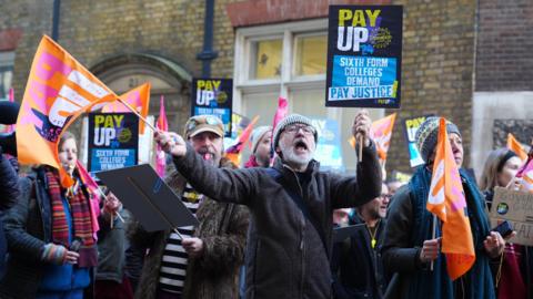 A group of men and women are standing with flags and banners saying pay up. One has a whistle in his mouth and another is shouting. 