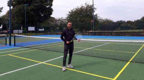 Councillor Mohammed Rafique stands on a tennis court in Leeds, while holding a tennis racket. The green court has yellow and white lines and is surrounded by trees and shrubs. 