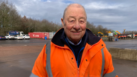 Mr Simpson is wearing an orange fluorescent jacket over a blue jumper and is stood outside in a work yard with some vehicles behind him