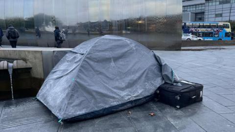 A grey tent on the pavement. People in thick coats walk past.