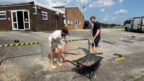 Two workmen work to clear debris from the ground outside a single-storey building. One man has a shovel and the other a broom. 