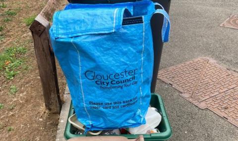 The image depicts a blue recycling sack resting atop a green recycling bin in an outdoor setting. The sack bears printed instructions from Gloucester City Council, encouraging residents to use it specifically for paper, card, and cardboard recycling as part of their city transformation initiative. Surrounding the bin are elements of a typical streetscape, including a pavement, patches of grass, a wooden post, and metal utility covers embedded in the pavement.