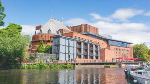 A large red-brick building on the riverside with grey clad sections. Boats can be seen on the right of the picture. There are parasols on a terrace overlooking the river. Pedestrians walk along a river path.