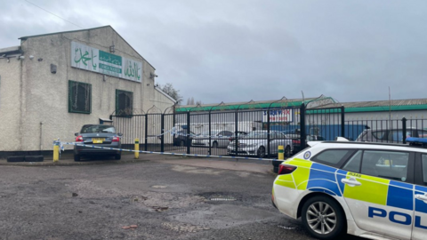 A large light brown building with a sloped roof and metal railings at the front and a police car parked in the foreground