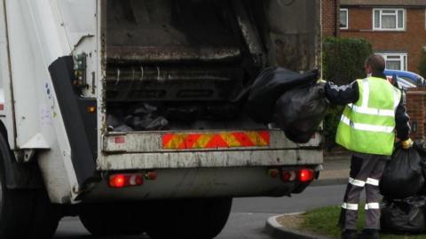 A refuse worker wearing a high-vis jacket puts black bin bags into the back of a white bin lorry.