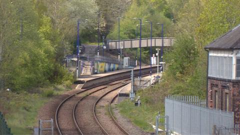 A view of the railway platform with some tracks leading up to it. There is a concrete bridge over the tracks and tall street lights lining either side. On the right there is a brick signal box. Trees surround the platform and tracks.