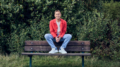 Man with blonde hair on a park bench with a hedge and grass around him.