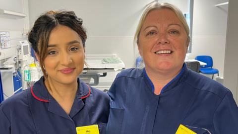Maj and Clare standing with their arms around each others' shoulders and looking into the camera and smiling. They're both wearing dark navy blue nurses' uniforms - Maj, on the left, has red borders on the collar, and Clare, on the right, has a lighter blue collar. They're both wearing bright yellow NHS name badges - but you can't quite make out what's written on them. They're standing in the entrance to one of the bed bays - a hospital bed is just visible beyond them, and the room has bright white walls and floors. 