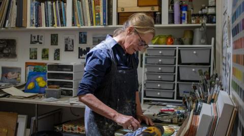 Woman working in blue overalls, standing with paintbrush in front of a canvas. There are colour charts on the wall and lots of drawers in the background.