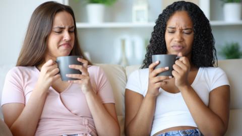 Two women looking distastefully in to their mugs whilst sat on the sofa