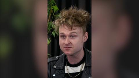 A man with spiky brown hair wearing a leather jacket and gold necklace