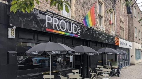 The facade of the former Proud Bar in Weston-super-Mare. Its sign features a rainbow motif and a rainbow flag hangs from a pole above the entrance. Chairs and tables are laid out on the pavement in front of the bar with sun umbrellas on them.