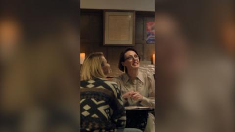Two women sitting across each other at a table in a restaurant, one is looking up talking to the server off-screen