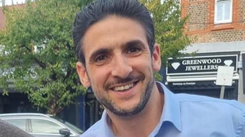 Yoni Finlay with black hair and beard wearing a blue shirt. He is standing in the street in front of a jewellers.