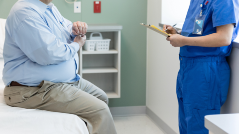 An overweight man sits on a bed in a clinical setting, in consultation with a medic wearing blue overalls. Their faces are cropped out of the picture