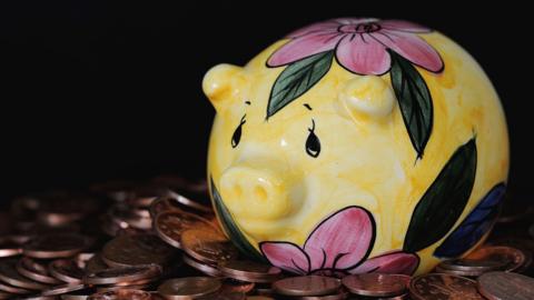 The image shows a yellow ceramic piggy bank with pink flowers and green leaves painted on it. It has black eyes and it is resting on a pile of coins.