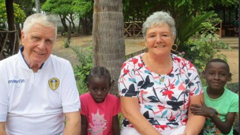 Dave and Sue Hayward - both with white short hair - sit smiling with two young African children in front of  tree and greenery in the African village. 