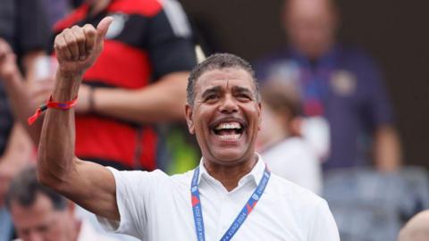 Chris Kamara waves from the stand before the UEFA EURO 2024 group stage match. He has short black hair and a wide smile.