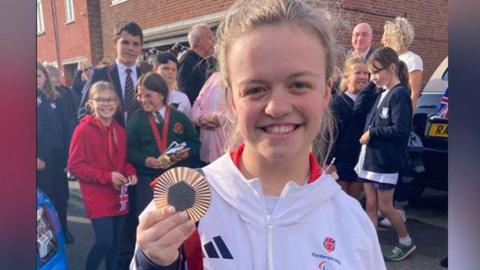 Champion Maisie Summers-Newton holding her medal after coming back from the Paralympics. She us smiling at the camera with her hair in a ponytail and is wearing a white Team GB hoodie.