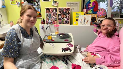 Patient Ruby, sitting up in her bed at Leeds Children's Hospital, and play specialist Megan Harpham