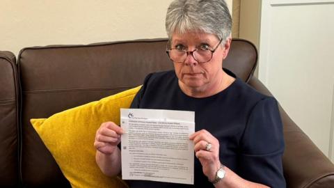 Alison Williams, a woman in her mid-60s with short, grey hair, holding a letter from the Civil Service Pensions Scheme informing her of delays to pension payments.