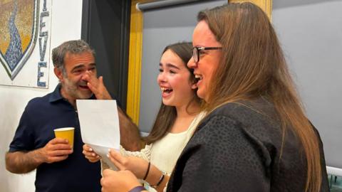 A student opening her results, looking happy with a man and woman next to her emotional and happy.