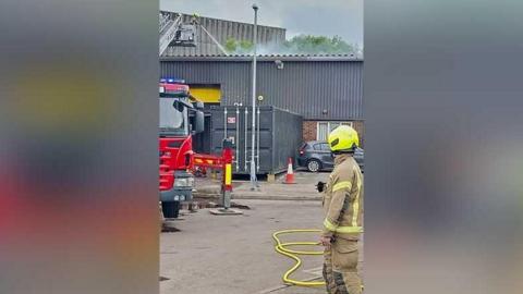 Smoke is seen rising from an industrial building as a firefighter in uniform watches on. There is a hose on the ground beside him.