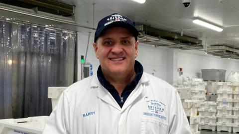 A man wearing a white shirt stands inside warehouse. He has his hands in his pocket and is wearing a blue cap. White boxes are stacked on top of each other around him. Strip lights are turned on. 