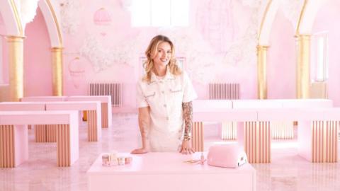A large room painted completely pink with pink tables and arches with gold columns. A woman is stood in the middle of the room with nail products smiling at the camera