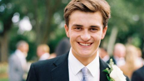 Henry Keith smiling. He appears to be at a wedding and is wearing a navy suit, white shirt and purple tie. He has a white button hole flower pinned to his jacket